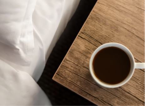 A white cup of black coffee sits on a wooden desk next to a bed.
